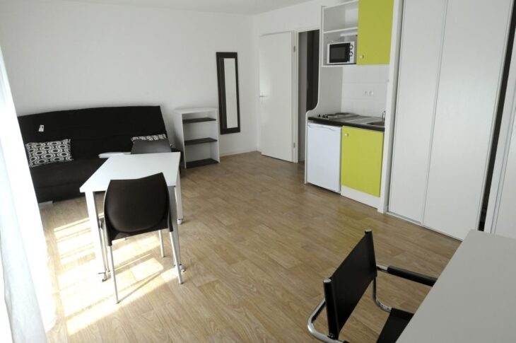 Studio meublé étudiant avec canapé-lit, coin cuisine intégré, miroir, étagère et placard blanc, vu en grand angle depuis une hauteur légère.