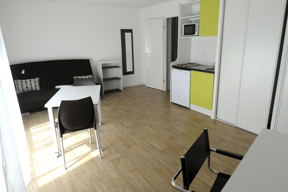 Studio meublé étudiant avec canapé-lit, coin cuisine intégré, miroir, étagère et placard blanc, vu en grand angle depuis une hauteur légère.